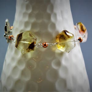 Pode incluir: Uma pulseira com pedras preciosas facetadas de cor &acirc;mbar e detalhes de borboletas prateadas. A pulseira &eacute; enfiada com uma corrente prateada e adornada com contas de cor cobre. As pedras preciosas t&ecirc;m uma forma geom&eacute;trica e s&atilde;o transl&uacute;cidas.