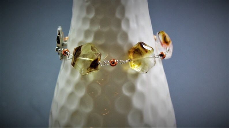 Pode incluir: Uma pulseira com pedras preciosas facetadas de cor &acirc;mbar e detalhes de borboletas prateadas. A pulseira &eacute; enfiada com uma corrente prateada e adornada com contas de cor cobre. As pedras preciosas t&ecirc;m uma forma geom&eacute;trica e s&atilde;o transl&uacute;cidas.