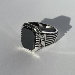 May include: A silver ring featuring a black rectangular stone and a Greek key design on the band.