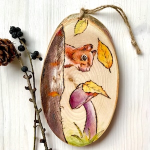Könnte beinhalten: Ein runder Holzschmuck mit einem gemalten Bild eines Eichhörnchens, das aus einem Baumstamm lugt und ein violettes Pilz mit gelben Blättern um sich herum betrachtet.