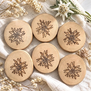 Pode incluir: Um conjunto de seis porta-copos circulares de madeira, cada um com uma ilustração escura gravada a fogo de uma flor edelweiss. Os porta-copos exibem padrões de veios de madeira natural e estão dispostos sobre um tecido claro e texturizado.