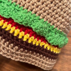 May include: Close-up of a crocheted hamburger made with brown, red, yellow and green yarn.