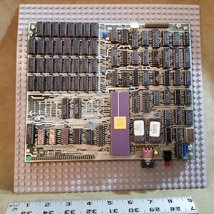 May include: A vintage computer circuit board with numerous black integrated circuits and a purple component. The board is mounted on a gray base with a ruler at the bottom, showing measurements in inches. The text "HSC INC C016" is visible.