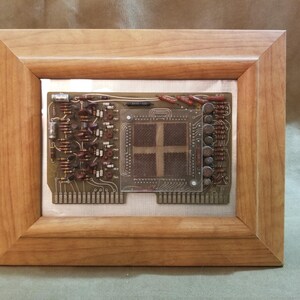 May include: A framed piece of artwork featuring a vintage circuit board with a central processor chip. The board is mounted on a silver background and framed in a light brown wood frame.