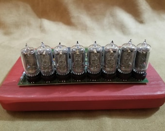 May include: A close-up of a circuit board with eight clear glass Nixie tubes, mounted on a red wooden base.