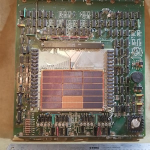 May include: A green circuit board with a large, rectangular, copper-colored chip in the center. The chip is surrounded by rows of smaller chips and wires. The board has a green background and black text that reads "7714".