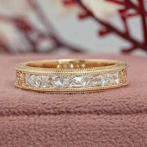 Puede incluir: Un anillo de banda dorado con piedras rectangulares y transparentes dispuestas en fila. El anillo tiene un dise&ntilde;o detallado alrededor de las piedras y a lo largo de los bordes. Se muestra sobre una superficie rosa, con un fondo floral borroso.