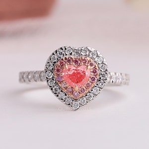 May include: A heart-shaped ring featuring a central pink stone surrounded by a halo of smaller pink stones, encased in a larger heart-shaped halo of clear white stones set in a metallic band with additional white stones along the shank.