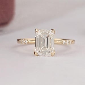 May include: A gold engagement ring with an emerald-cut diamond center stone. The ring features smaller accent stones on the band. The ring is set against a soft, neutral background, with a hint of a pink box in the background.