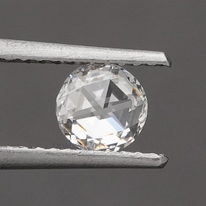 K&ouml;nnte beinhalten: Ein runder, klarer, facettierter Edelstein, der von einer silberfarbenen Metallpinzette gehalten wird. Der Stein hat einen Rosenschliff mit flacher Unterseite und einer gew&ouml;lbten, facettierten Oberseite, die Licht reflektiert.