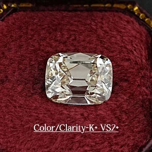 May include: A cushion-cut diamond with a light hue, set in a burgundy velvet jewelry box. The diamond's facets reflect light, creating a brilliant sparkle. Text on the box reads "Color/Clarity-K+ VS2+".