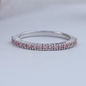May include: A silver band ring with a continuous row of small, pink gemstones. The ring is set against a soft, white background, with a blurred object in the background. The ring is a delicate piece of jewelry.