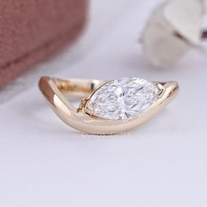 May include: A gold ring with a marquise-cut diamond. The diamond is set in a curved, open-style band. The ring is displayed on a white surface, with a blurred background.