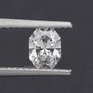 May include: A clear, faceted diamond held by silver tweezers. The diamond has an octagonal shape with multiple facets, reflecting light. The background is a dark gray, providing contrast to the bright gemstone. The text "OUROS JEWELS" is visible.