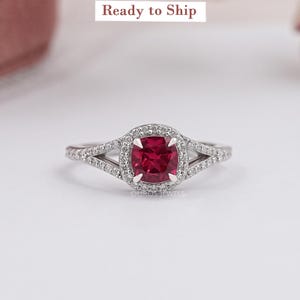 May include: A silver ring with a square, deep red gemstone at the center, surrounded by a halo of smaller, clear stones. The band splits into two on each side, also adorned with small, clear stones. The text "Ready to Ship" is visible.