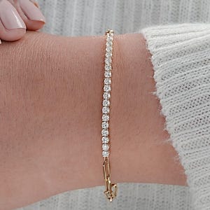 May include: A gold bracelet with a row of small, clear gemstones. The bracelet is clasped and worn on a wrist, with a light-colored sweater in the background. The gemstones are evenly spaced and reflect light.