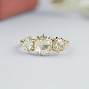 May include: A gold ring featuring three round, faceted, clear stones set in a horizontal row. The center stone is larger than the two flanking stones, and each is held in place by small metal prongs on a thin, polished gold band.