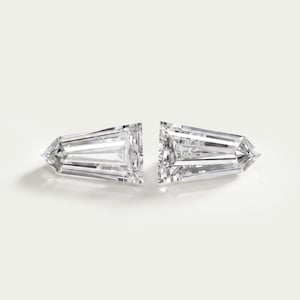 May include: Two clear, colorless, faceted gemstones with a tapered baguette cut. Each stone features a flat, wide top edge and a pointed bottom edge, with multiple internal facets reflecting light to create a brilliant, transparent appearance.