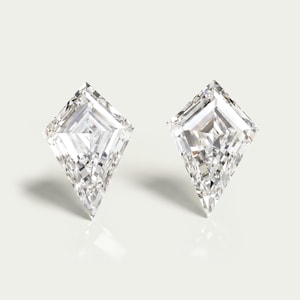 May include: A pair of clear, colorless, kite-shaped faceted gemstones. Each stone features a sharp, pointed bottom and a wider, angular top section with multiple internal facets that reflect light, creating a brilliant, sparkling appearance.