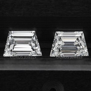 Puede incluir: Dos diamantes transparentes de talla esmeralda, de forma cuadrada, sobre un fondo negro. Los diamantes están colocados en una bandeja negra con las palabras "Ouro's Jewels" impresas debajo de cada diamante.