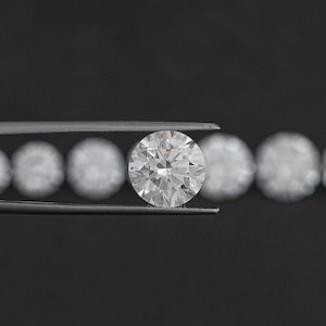 May include: A round, clear diamond gemstone held in tweezers. The diamond is surrounded by a row of smaller, round diamonds.