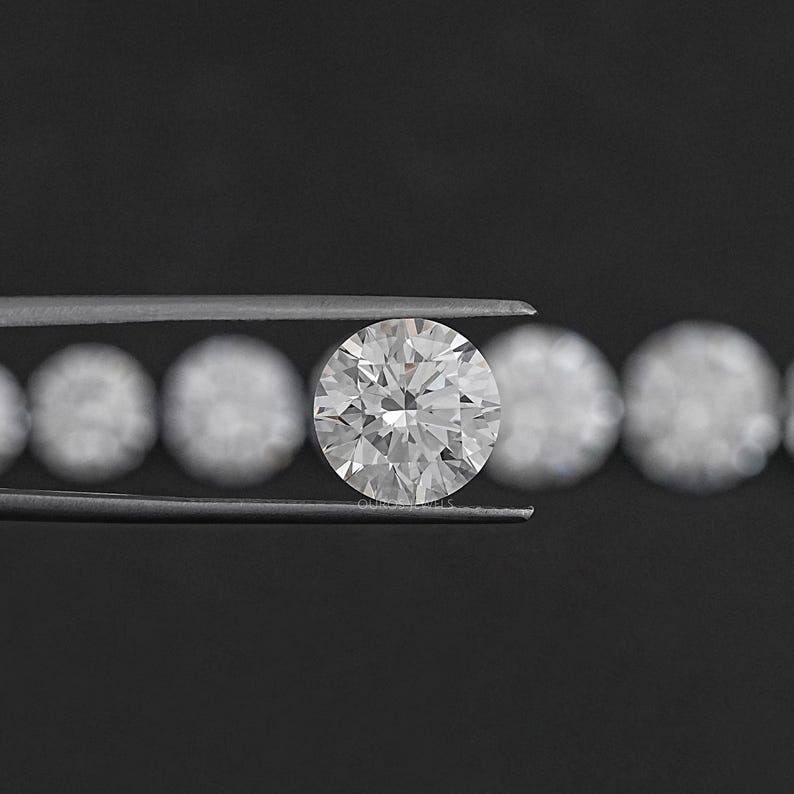 May include: A round, clear diamond gemstone held in tweezers. The diamond is surrounded by a row of smaller, round diamonds.