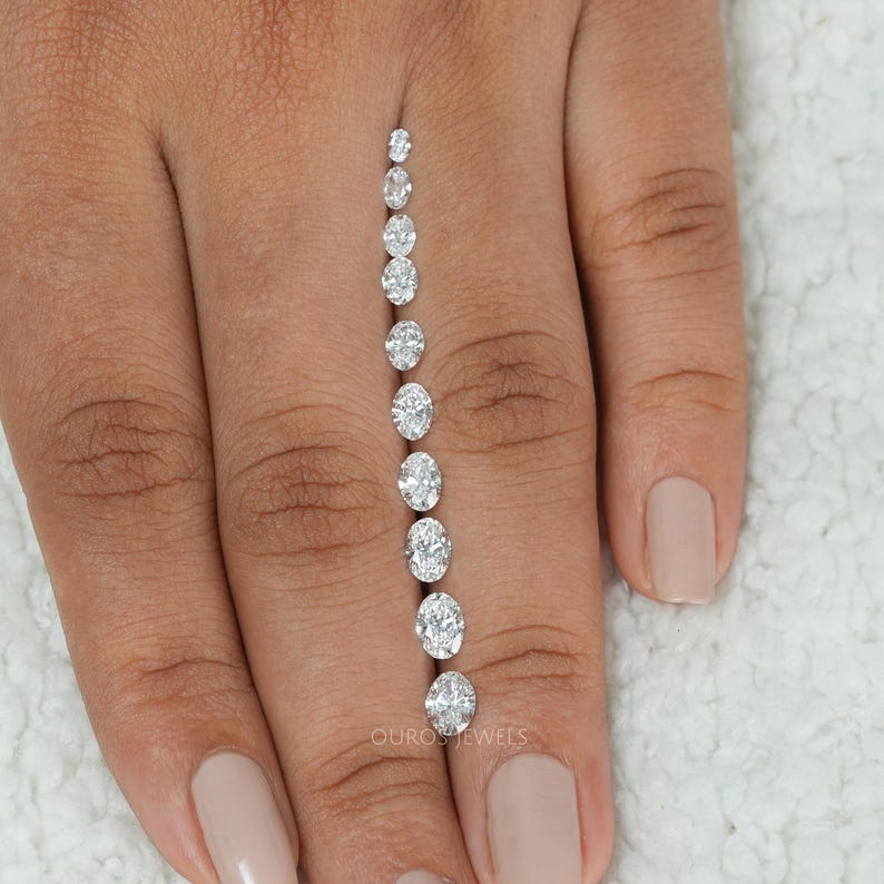 May include: A row of oval-shaped diamonds of varying sizes, arranged from smallest to largest, on a person's finger.