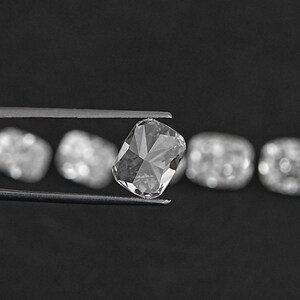 K&ouml;nnte beinhalten: Ein klarer, facettierter, rechteckiger Diamantenstein, der in einer Pinzette gehalten wird. Der Diamant ist von anderen &auml;hnlichen Edelsteinen umgeben, die sich alle auf einem schwarzen Hintergrund befinden.