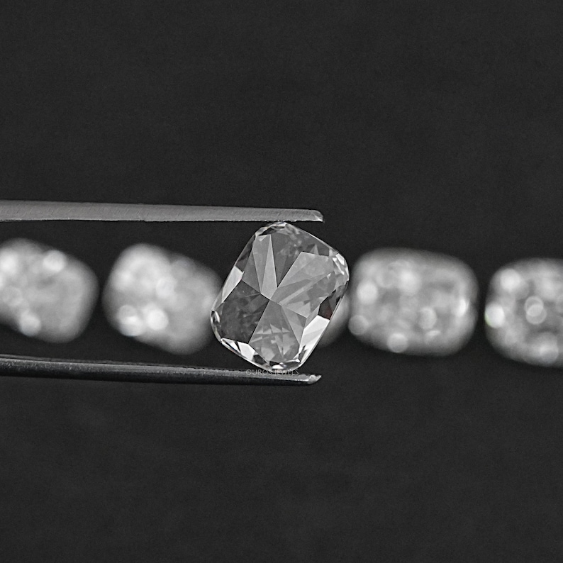K&ouml;nnte beinhalten: Ein klarer, facettierter, rechteckiger Diamantenstein, der in einer Pinzette gehalten wird. Der Diamant ist von anderen &auml;hnlichen Edelsteinen umgeben, die sich alle auf einem schwarzen Hintergrund befinden.