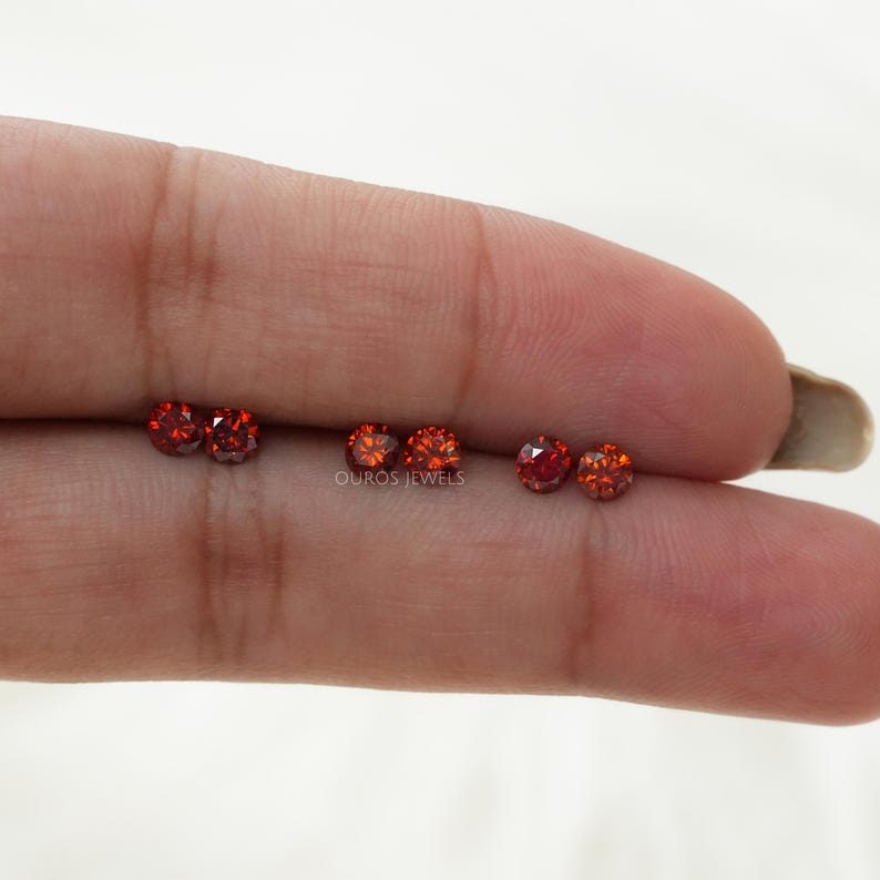 Puede incluir: Cinco piedras preciosas redondas de color rojo anaranjado en un dedo. Las piedras preciosas son peque&ntilde;as y tienen un corte facetado. El texto "OUROS JEWELS" es visible debajo de las piedras preciosas.