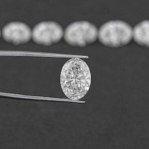 Pu&ograve; includere: Un diamante ovale trasparente tenuto in una pinza su uno sfondo nero. Il diamante &egrave; circondato da una fila di diamanti pi&ugrave; piccoli.