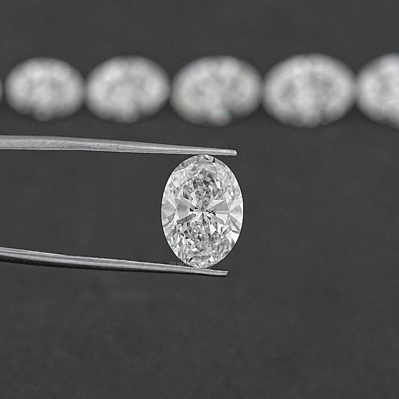 Pu&ograve; includere: Un diamante ovale trasparente tenuto in una pinza su uno sfondo nero. Il diamante &egrave; circondato da una fila di diamanti pi&ugrave; piccoli.