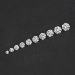 May include: A row of ten round, clear, faceted gemstones of varying sizes, arranged from smallest to largest on a black surface.