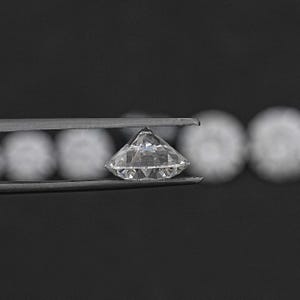 May include: A clear, round diamond gemstone held in tweezers. The diamond is faceted and reflects light.