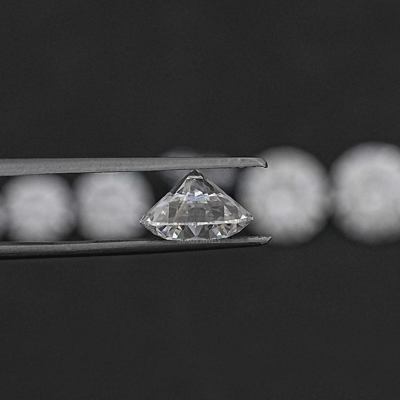 May include: A clear, round diamond gemstone held in tweezers. The diamond is faceted and reflects light.