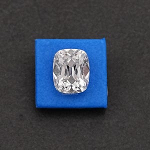 May include: A large, clear cushion-cut diamond with brilliant facets, set on a square of blue fabric. The diamond reflects light, showcasing its clarity and sparkle. The background is a dark, neutral color.