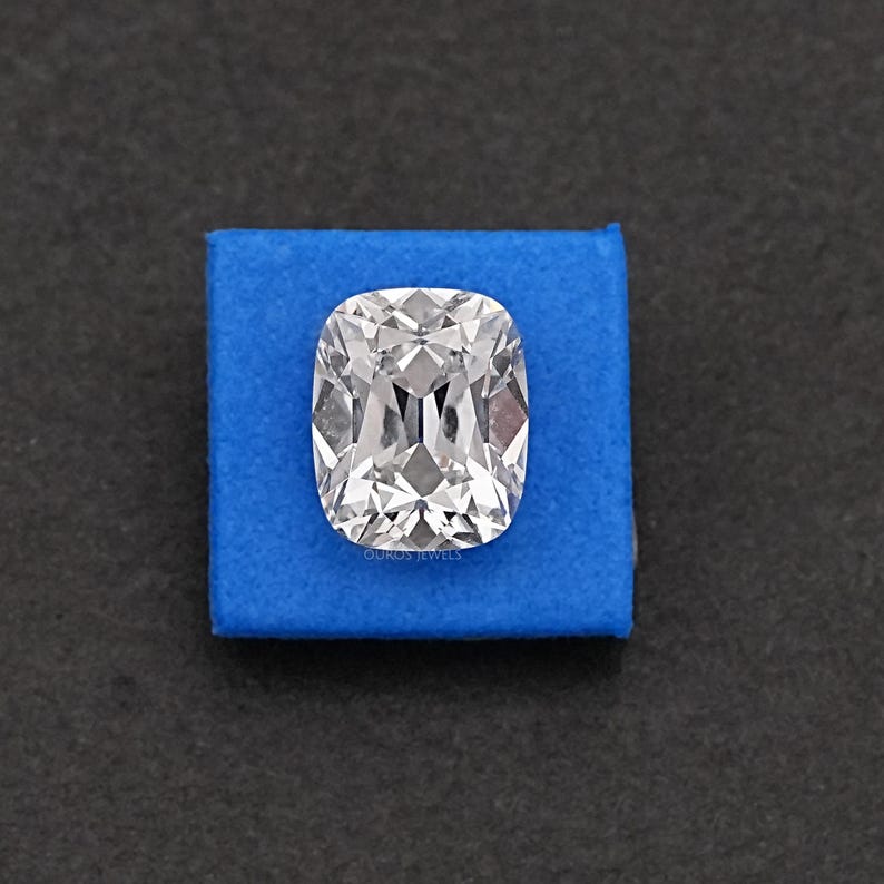 May include: A large, clear cushion-cut diamond with brilliant facets, set on a square of blue fabric. The diamond reflects light, showcasing its clarity and sparkle. The background is a dark, neutral color.