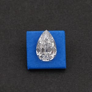 May include: A pear-shaped, clear diamond gemstone on a blue velvet cushion. The diamond is faceted and reflects light.