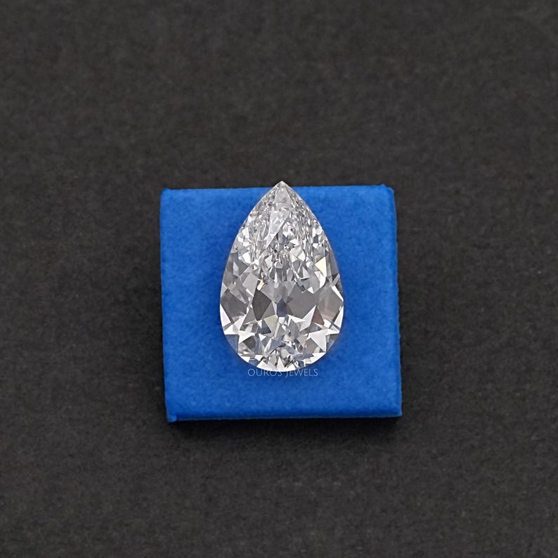 May include: A pear-shaped, clear diamond gemstone on a blue velvet cushion. The diamond is faceted and reflects light.