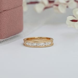 Puede incluir: Un anillo de banda de oro con una serie de piedras preciosas claras de corte cuadrado. El anillo tiene un delicado dise&ntilde;o de borde con cuentas. El anillo se muestra sobre una superficie blanca, con una caja de joyer&iacute;a rosa en el fondo.