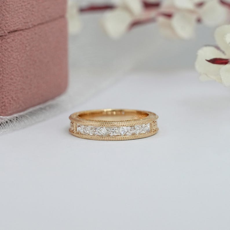 Puede incluir: Un anillo de banda de oro con una serie de piedras preciosas claras de corte cuadrado. El anillo tiene un delicado dise&ntilde;o de borde con cuentas. El anillo se muestra sobre una superficie blanca, con una caja de joyer&iacute;a rosa en el fondo.