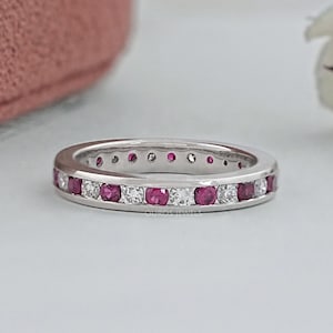 May include: A silver-toned metal eternity band featuring an alternating pattern of round-cut, deep pink rubies and clear, sparkling white diamonds set in a channel setting. The ring displays a polished finish and a consistent, circular design.