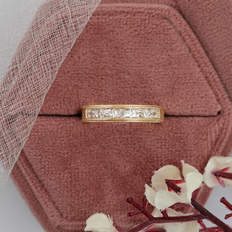 Puede incluir: Un anillo de banda de oro con una fila de piedras claras de corte cuadrado. El anillo est&aacute; sobre una superficie de terciopelo rosa. El borde del anillo es detallado y texturizado. El fondo incluye tela blanca transparente y flores.