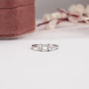 May include: A silver-toned ring featuring a central rectangular setting designed to hold a stone, flanked by two small, clear, heart-shaped stones on either side of the band. The metal band is polished and smooth, showcasing a clean, metallic finish.
