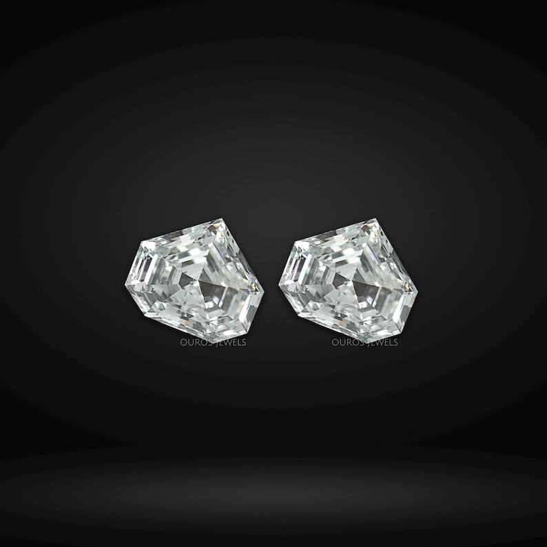 May include: Two loose, colorless, pentagon-shaped faceted gemstones with a brilliant cut. Each stone features a symmetrical arrangement of triangular and trapezoidal facets that reflect light. The text OUROS JEWELS is printed in small white letters below each stone.
