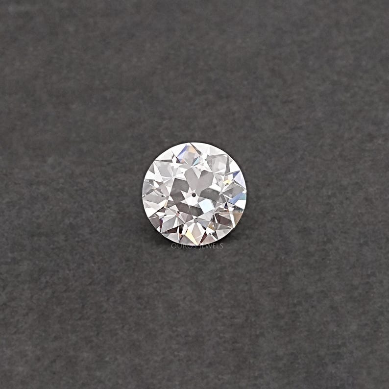 May include: A round, clear, faceted diamond gemstone on a gray background.