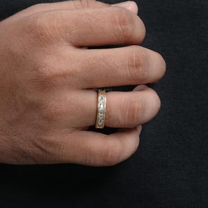 Puede incluir: Un anillo de banda de oro con piedras preciosas claras dispuestas en fila. El anillo est&aacute; en el dedo de una persona. El anillo tiene un dise&ntilde;o texturizado a lo largo de los bordes. El fondo es oscuro.
