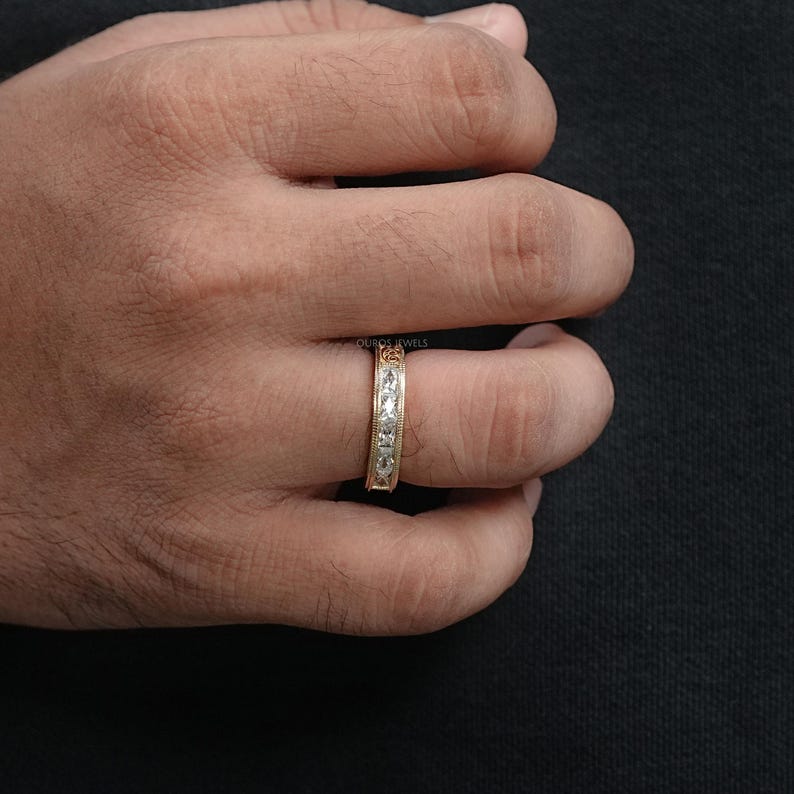 Puede incluir: Un anillo de banda de oro con piedras preciosas claras dispuestas en fila. El anillo est&aacute; en el dedo de una persona. El anillo tiene un dise&ntilde;o texturizado a lo largo de los bordes. El fondo es oscuro.