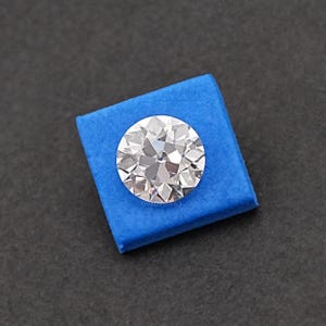 May include: A clear round diamond gemstone on a blue velvet background. The diamond has many facets and is sparkling.