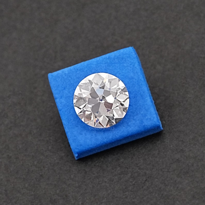May include: A clear round diamond gemstone on a blue velvet background. The diamond has many facets and is sparkling.